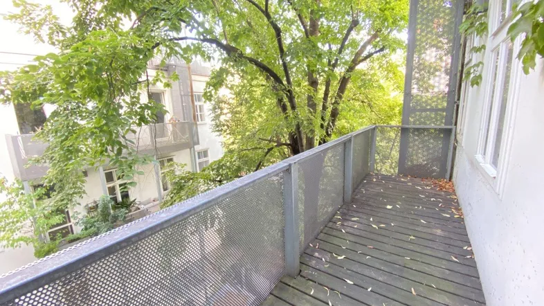 Geräumiger Balkon mit Holzboden und Metallgeländer, umgeben von üppigem Grün und Blick auf Nachbargebäude.