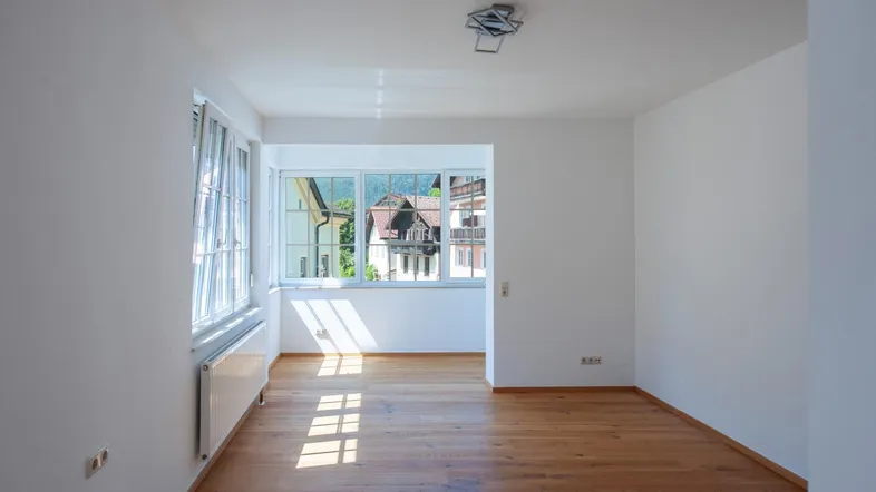 Helles, leeres Zimmer mit Holzboden und großen Fenstern, die einen Blick ins Grüne bieten.