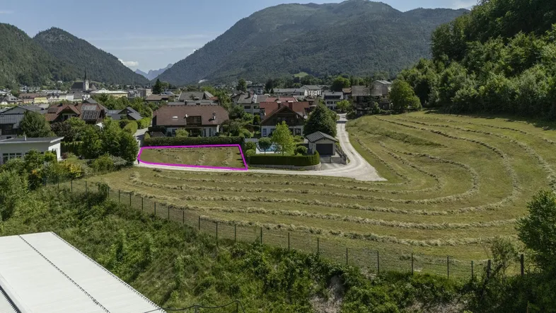 Luftaufnahme eines Baugrundstücks mit umliegenden Häusern, Feldern und Bergpanorama.
