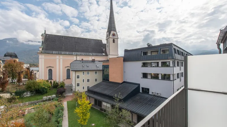 Blick vom Balkon auf eine Kirche, umliegende Gebäude und eine grüne Gartenanlage mit Bergen im Hintergrund.