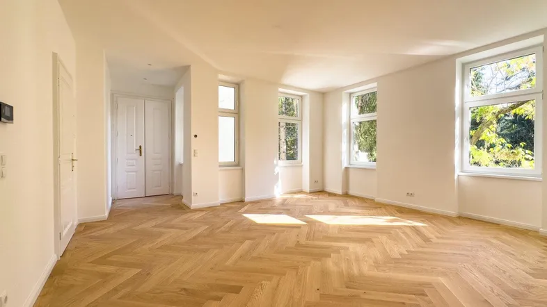 Helles, geräumiges Zimmer mit Fischgrätparkettboden, weißen Wänden und großen Fenstern, die viel Licht hereinlassen.