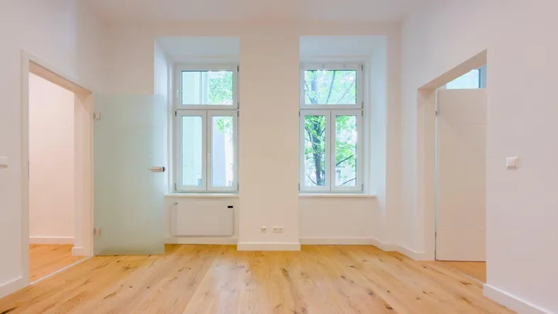 Helles, leeres Zimmer mit Holzboden, weißen Wänden und zwei Fenstern mit Blick ins Grüne.