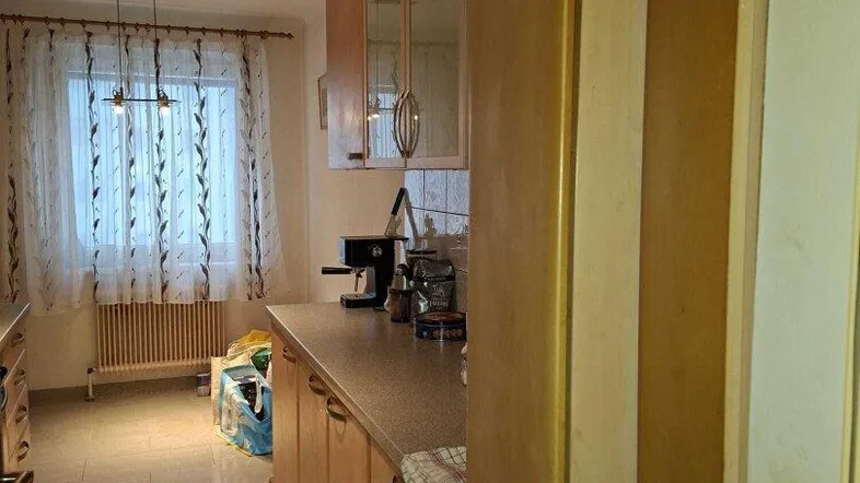 Helle Küche mit Holzschränken, Fliesenboden und Fenster mit Vorhängen.