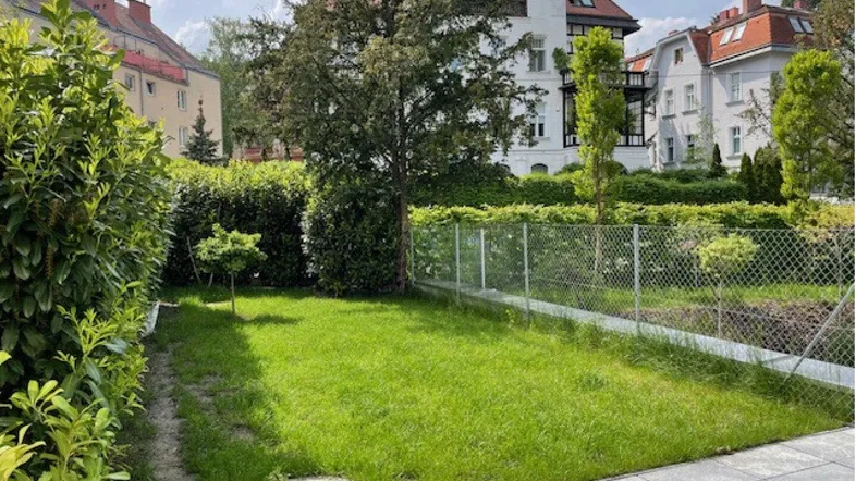 Gepflegter Gartenbereich mit Rasenfläche, Hecke und gepflasterter Terrasse hinter dem Gebäude.