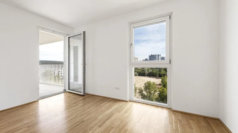 Helles Zimmer mit Parkettboden, großem Fenster und Zugang zu einem Balkon.