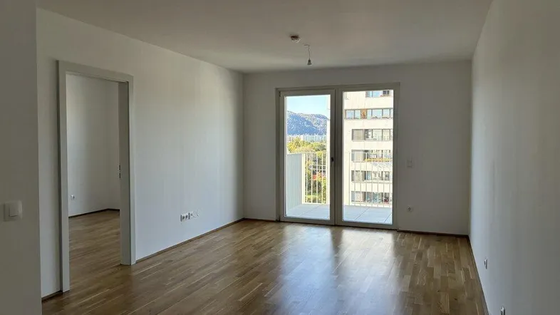 Helles Zimmer mit Parkettboden und Zugang zu einem Balkon, bietet einen Blick auf die umliegenden Gebäude und Berge.