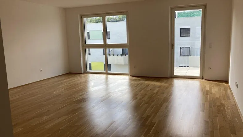 Helles, leeres Wohnzimmer mit Parkettboden und großen Fenstern sowie Zugang zu einem Balkon.