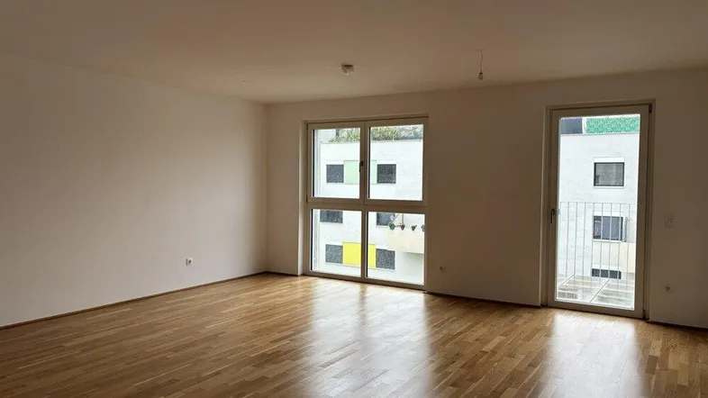 Helles, leeres Wohnzimmer mit Parkettboden und Zugang zu einem kleinen Balkon.