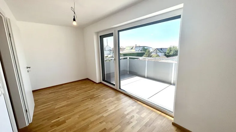 Heller Raum mit Parkettboden und großem Fenster, das Zugang zu einem Balkon bietet.