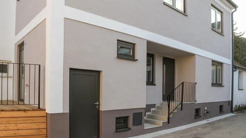 Außenansicht des Gebäudes mit moderner Fassade, Holzterrasse und Zugang zum Eingangsbereich.