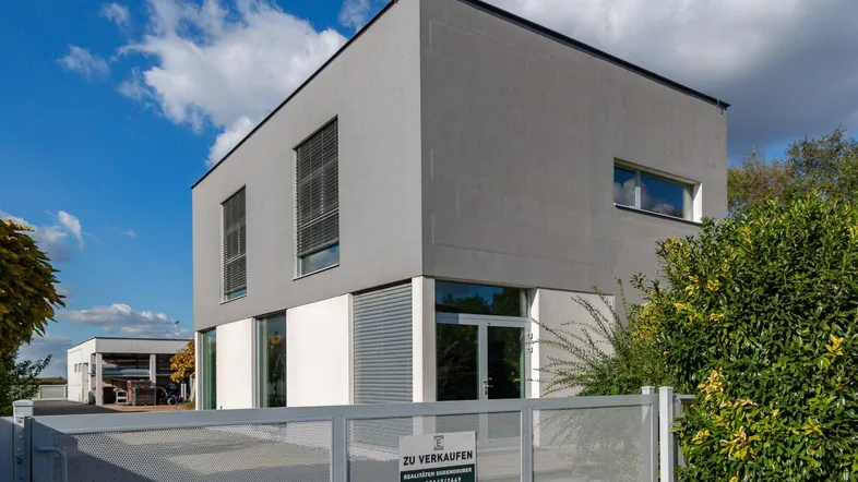 Modernes Gebäude mit grauer Fassade, großen Fenstern und einem Zaun im Vordergrund.