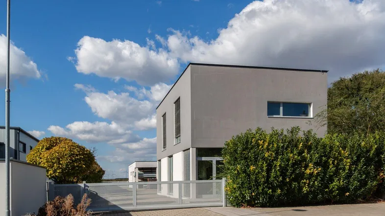 Modernes graues Gebäude mit klaren Linien und großen Fenstern, umgeben von grünen Büschen unter blauem Himmel.