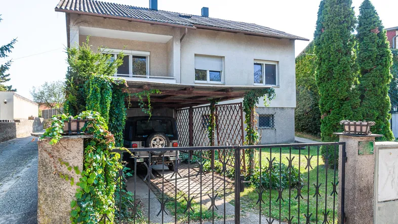 Einfamilienhaus mit Carport und Vorgarten, umgeben von Bäumen und einem Zaun.