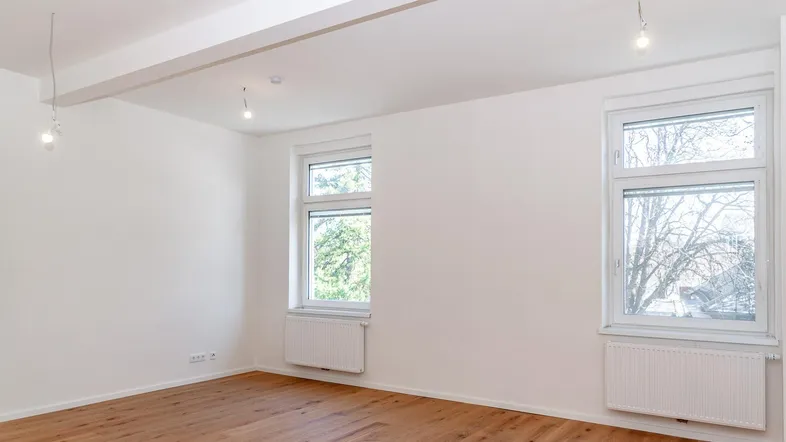 Helles, leeres Zimmer mit Holzboden, weißen Wänden und zwei Fenstern, bereit zum Einzug.