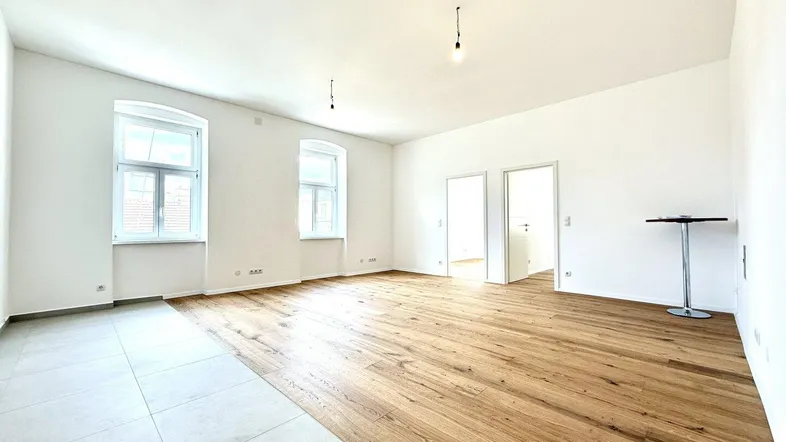 Geräumiger, leerer Wohnbereich mit Holzboden und Fliesen, großen Fenstern und viel Tageslicht.