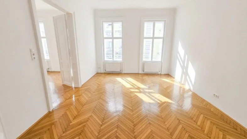Helles Zimmer mit Fischgrätparkett, weißen Wänden und zwei Fenstern, das viel Tageslicht hereinlässt.