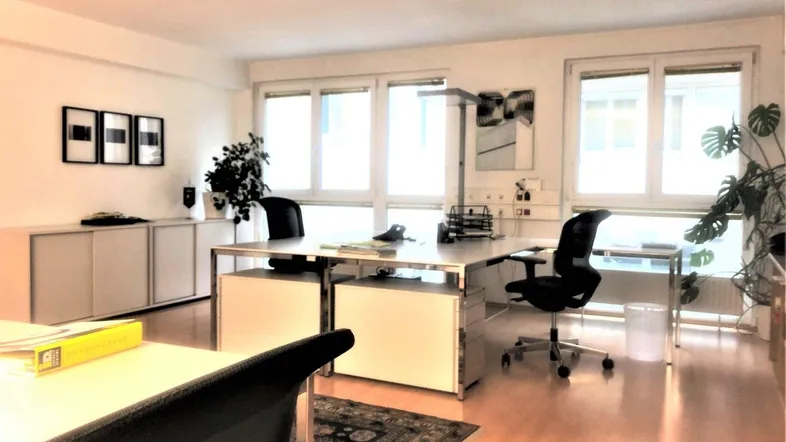 Helles Büro mit mehreren Arbeitsplätzen, großen Fenstern und Holzfußboden, ausgestattet mit Büromöbeln.