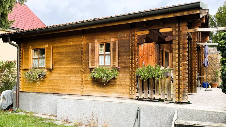 Seitenansicht des Holzhauses mit traditionellen Fensterläden und einem kleinen Gartenbereich.