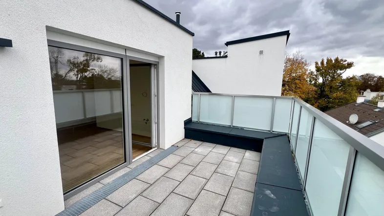 Moderne Dachterrasse mit Fliesenboden, Glasgeländer und Zugang zu einem hellen Innenraum.