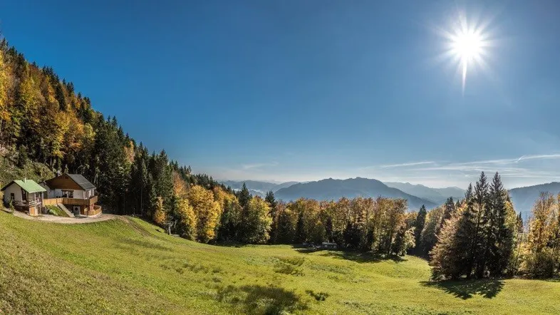 Hütte in alpiner Landschaft mit grünen Wiesen und herbstlichen Bäumen unter strahlendem Sonnenschein.