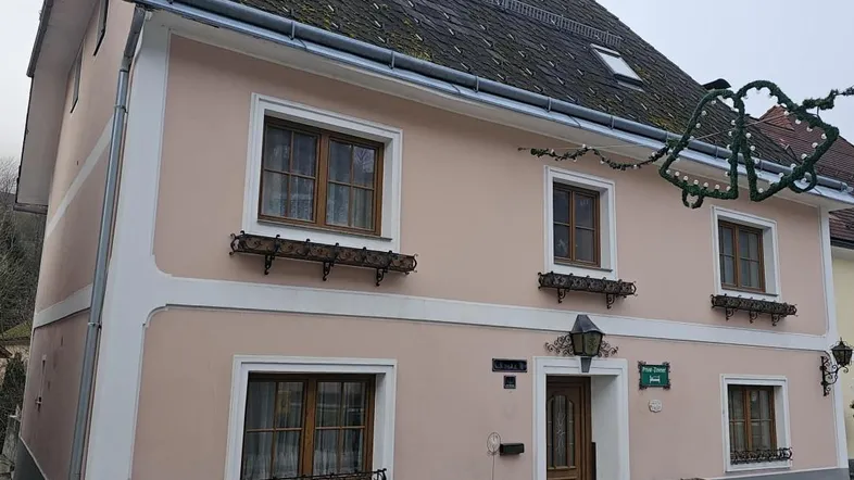 Traditionelles Haus mit pastellrosa Fassade, Holzfenstern und einem Satteldach in ländlicher Umgebung.