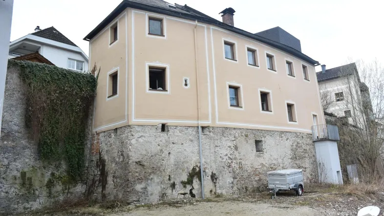 Historisches Gebäude mit steinerner Basis und hellbeiger Fassade, umgeben von älterer Bausubstanz.