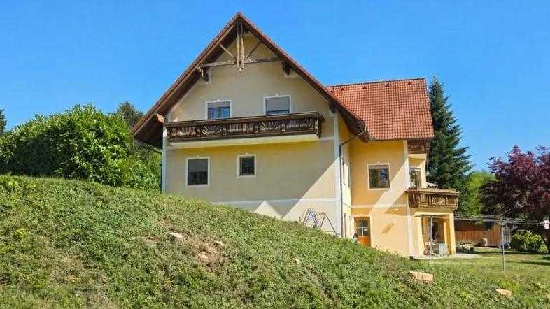 Freistehendes Einfamilienhaus mit gelber Fassade, roten Ziegeldächern und traditionellen Holzbalkonen, eingebettet in eine grüne Hügellandschaft.