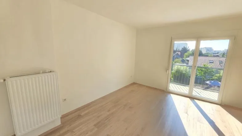 Helles Zimmer mit Laminatboden, weiß gestrichenen Wänden und Zugang zu einem Balkon mit Ausblick.