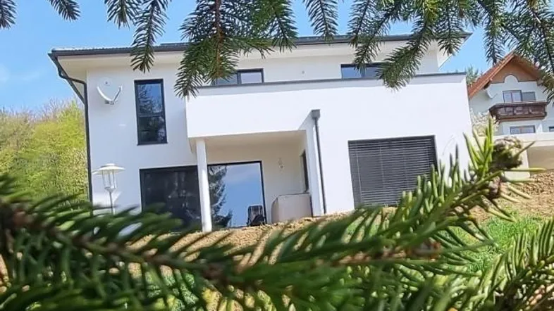 Modernes Einfamilienhaus mit weißer Fassade und großen Fenstern, umgeben von Natur.