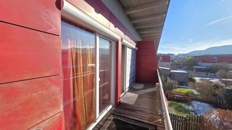 Sonniger Balkon mit Holzboden, roter Fassade und weitem Blick auf die Umgebung.