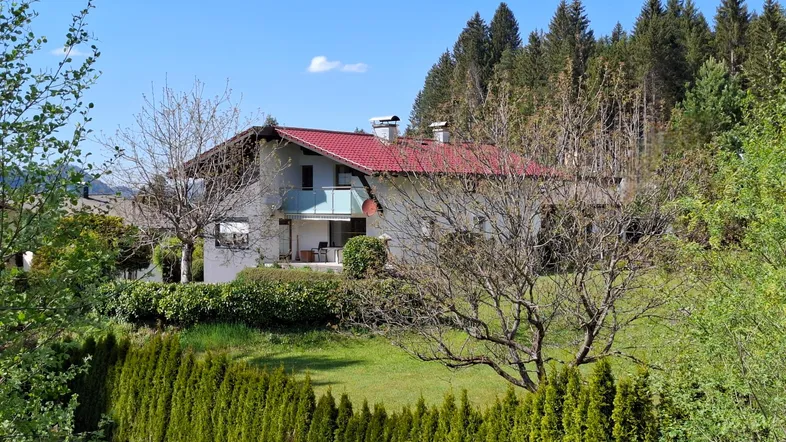 Freistehendes Haus mit rotem Dach, umgeben von üppigem Grün und hohen Bäumen unter blauem Himmel.