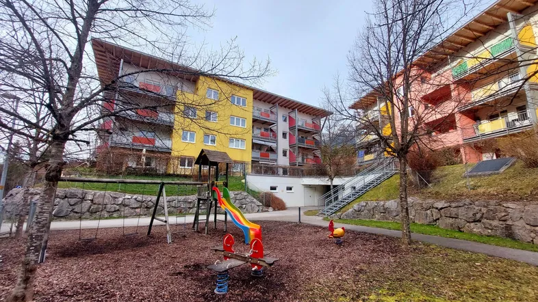 Bunte Mehrfamilienhäuser mit Spielplatz im Vordergrund, umgeben von Bäumen und Grünflächen.