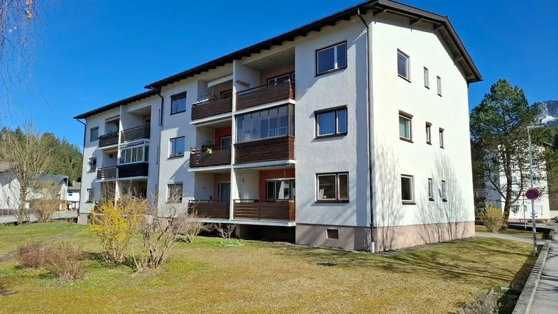 Mehrfamilienhaus mit mehreren Balkonen und gepflegter Grünfläche im Vordergrund unter blauem Himmel.