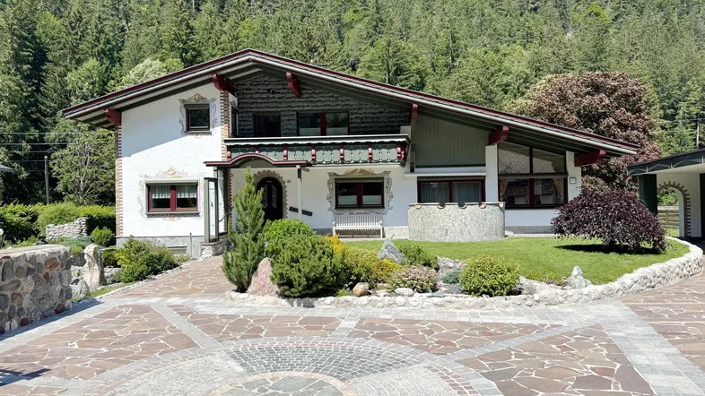 Traditionelles Alpenhaus mit gepflasterter Auffahrt, gepflegtem Vorgarten und bewaldetem Berghintergrund.