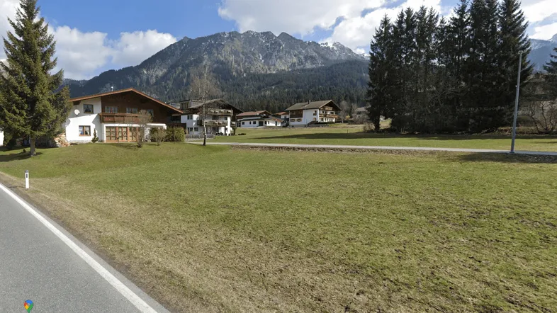 Häuser in einer alpinen Landschaft mit schneebedeckten Bergen im Hintergrund und grünen Wiesen im Vordergrund.