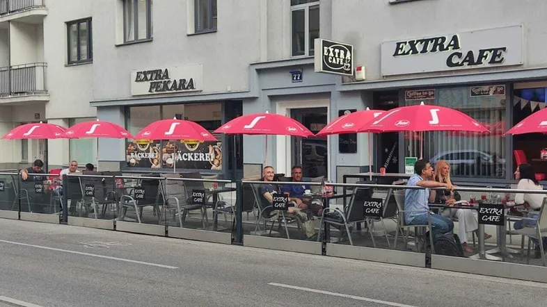 Straßencafé mit roten Sonnenschirmen und Glasabtrennung, belebt mit Gästen.