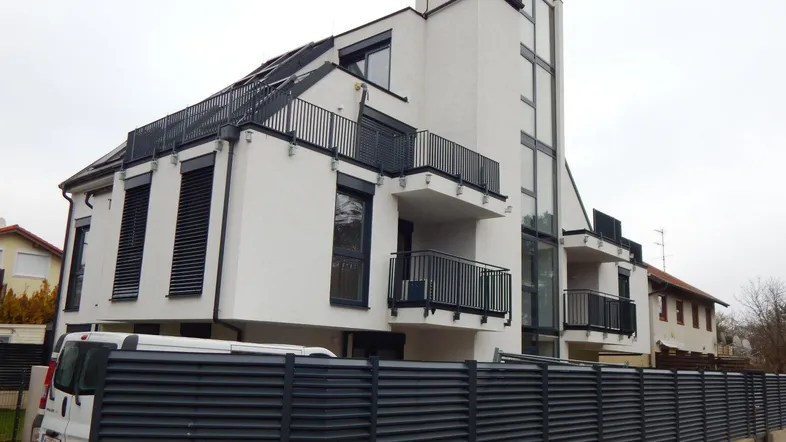 Modernes Mehrfamilienhaus mit weißer Fassade, Balkonen und großen Fenstern unter bewölktem Himmel.