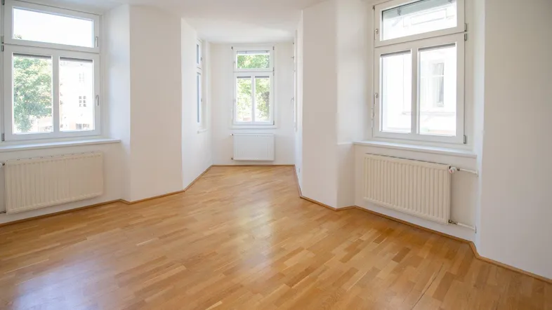 Heller, leerer Raum mit Parkettboden, weißen Wänden und mehreren Fenstern, die viel Tageslicht hereinlassen.