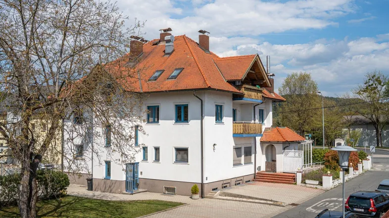 Großes Wohngebäude mit traditionellem Ziegeldach, blauen Fensterrahmen und gepflastertem Vorplatz.