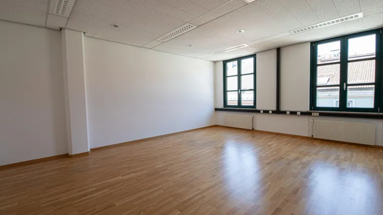 Geräumiges, leeres Büro mit hellem Parkettboden und großen Fenstern, die viel Tageslicht hereinlassen.
