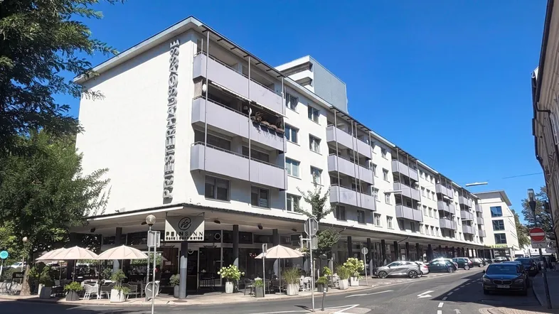 Mehrstöckiges Wohn- und Geschäftshaus mit Balkonen und Straßencafé an einem sonnigen Tag.