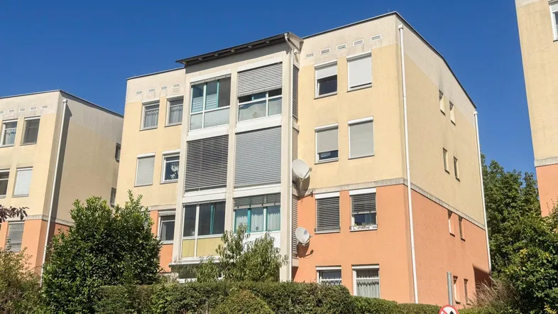 Mehrfamilienhaus mit mehreren Etagen, Balkonen und Rollläden unter blauem Himmel.