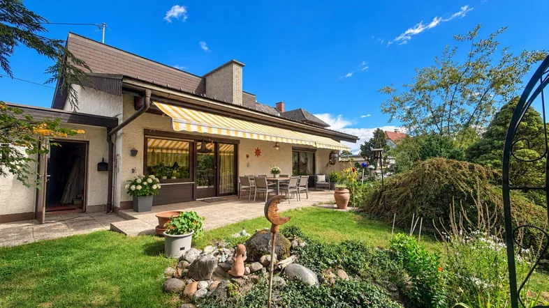 Geräumige Terrasse mit Markise und Gartenmöbeln, umgeben von einem gepflegten Garten unter blauem Himmel.