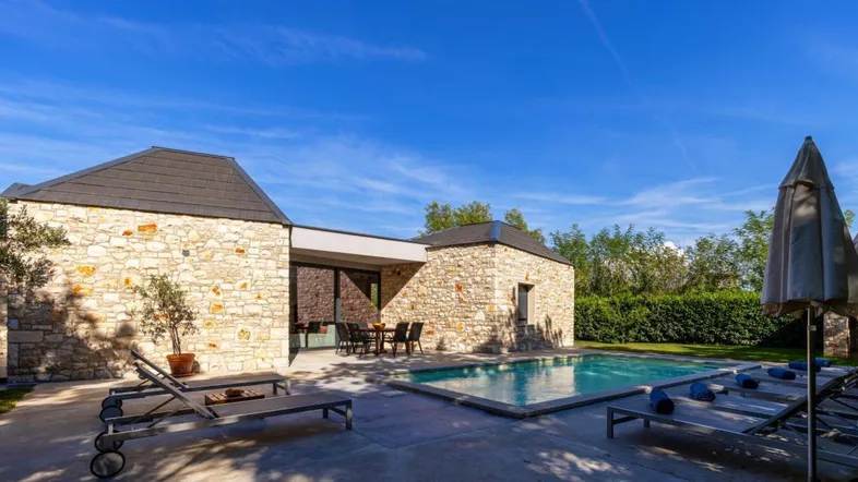 Außenansicht der Villa mit großem Poolbereich, Sonnenliegen und Essbereich im Freien unter blauem Himmel.