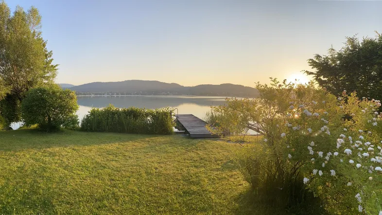 Idyllischer Gartenbereich mit direktem Seezugang und Steg bei Sonnenuntergang, umgeben von grüner Natur und Bergen.