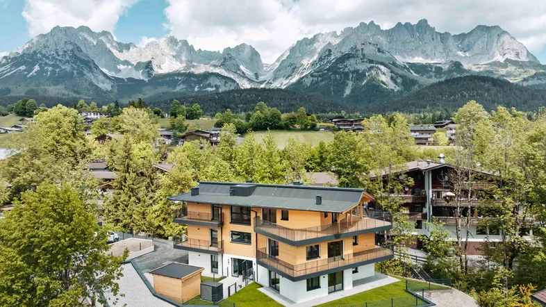 Modernes Apartmentgebäude mit Holzfassade und Balkonen vor beeindruckender Bergkulisse in grüner Umgebung.
