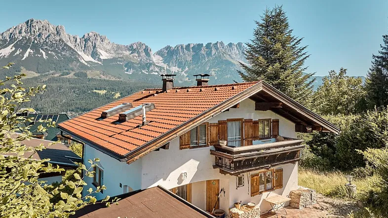 Traditionelles Haus mit rotem Ziegeldach und Balkon, umgeben von grüner Natur und majestätischen Bergen.