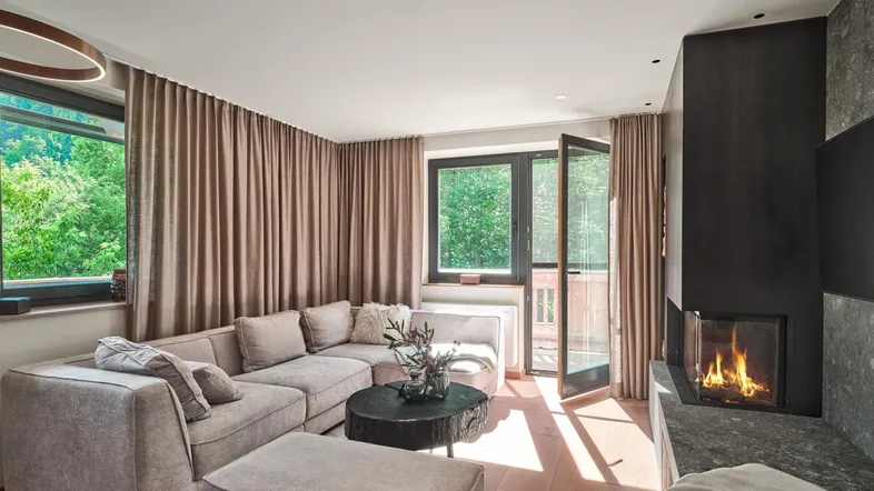 Gemütliches Wohnzimmer mit großem Ecksofa, modernem Kamin und Zugang zu einem Balkon mit Grünblick.