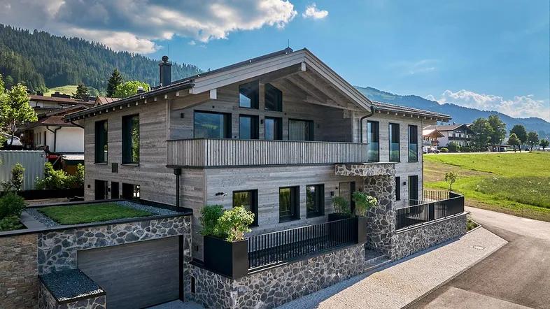 Modernes Chalet mit Holzfassade, Steinelementen und Bergblick unter blauem Himmel.