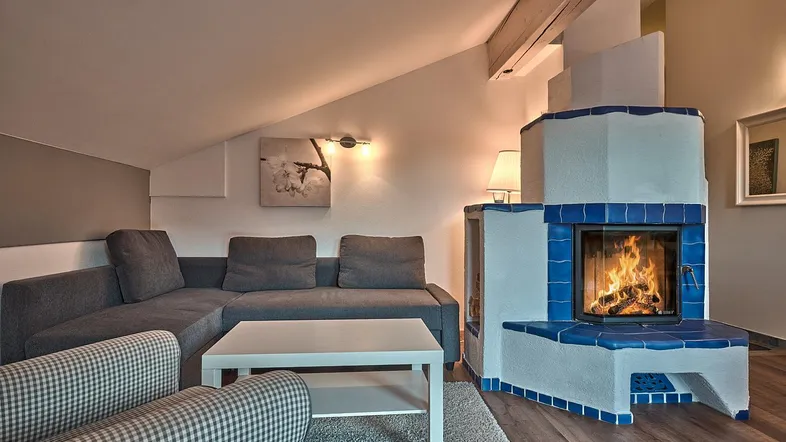 Gemütliches Wohnzimmer im Dachgeschoss mit Ecksofa, Teppich und einem traditionellen Kachelofen mit blauen Akzenten.